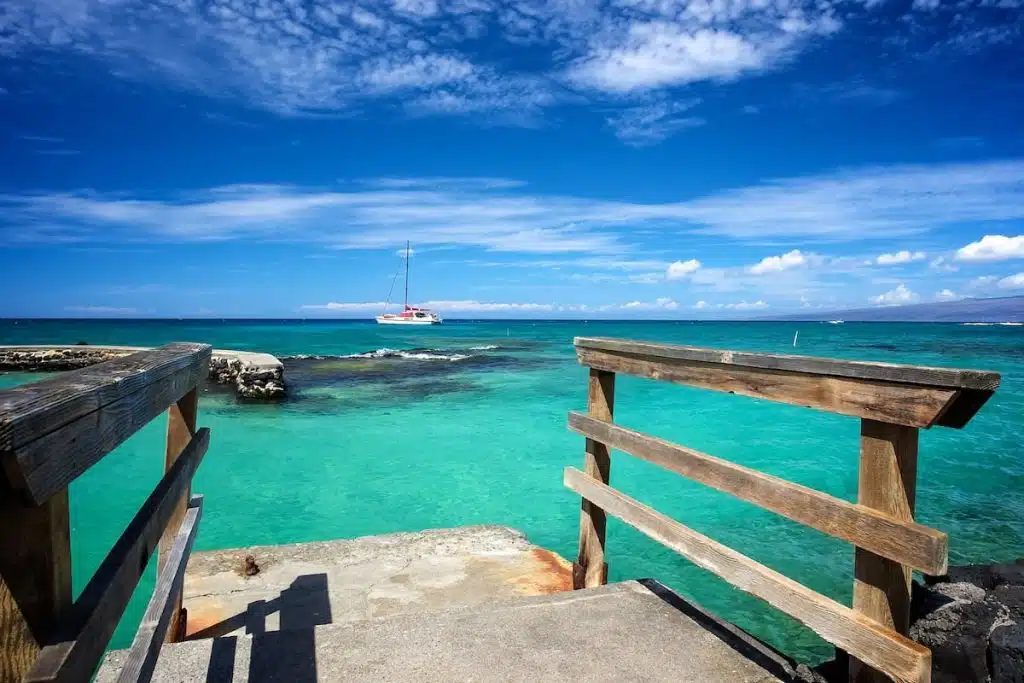 A wooden railing leads down to a turquoise sea, with a distant sailboat on the horizon under a blue sky with scattered clouds.