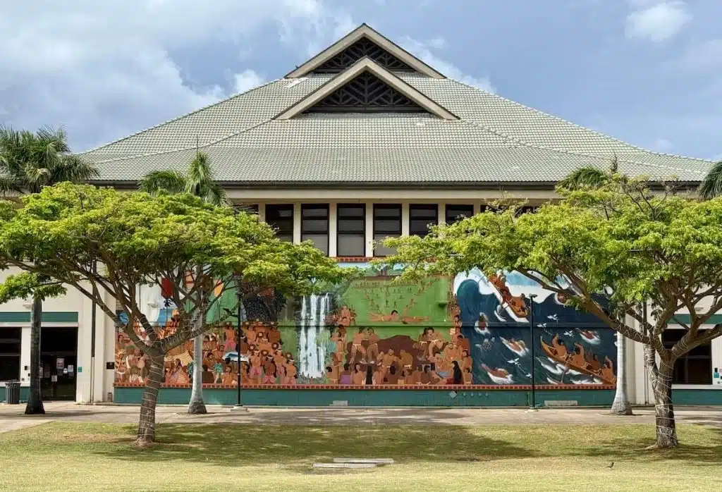 A large building with a prominent gray roof is adorned with a colorful mural depicting cultural scenes, including people and nature, along the front wall. Lush green trees partially obscure the artwork, while the sky is filled with light clouds.