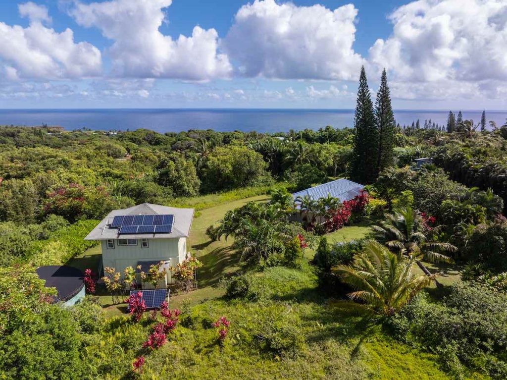 June 2026 Maui Statistics and Market Update 1 This photo shows an aerial image of an off the grid home with a detached garage and ag office set in a lush landscape of palm trees, cook island palms, vibrant red ti plants. A vibrant blue ocean is close by. Fluffy white clouds carried by the trade winds bring occasional showers.
