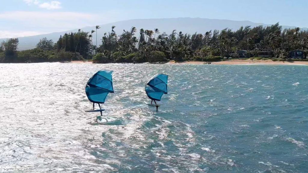 two people wing foiling off the coast of Spreckelsville on Maui's North Shore. Haleakala volcano can be seen in the distance behind the palm tree lined beach. 