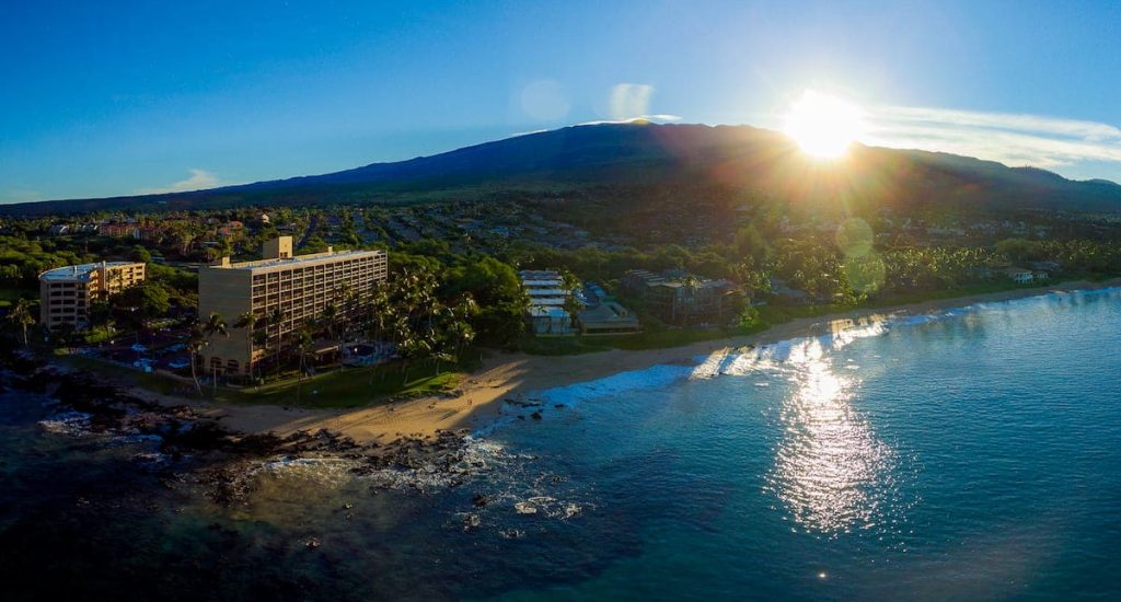Aerial view of a coastal resort area with sandy beaches, palm trees, and buildings, set against a backdrop of mountains and a sunrise or sunset over the ocean.